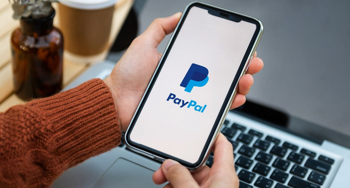 Man holding smartphone with open PayPal app on laptop background