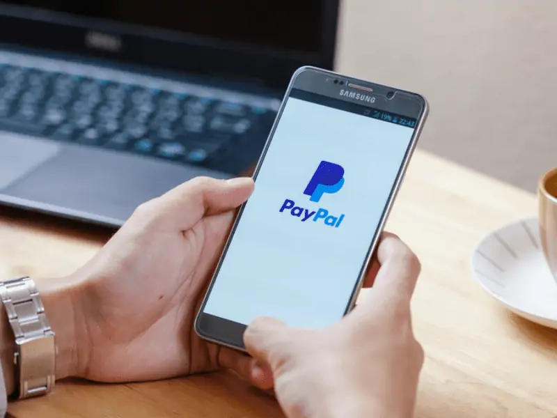 A man is holding a smartphone with a PayPal icon on the background of a laptop and a cup