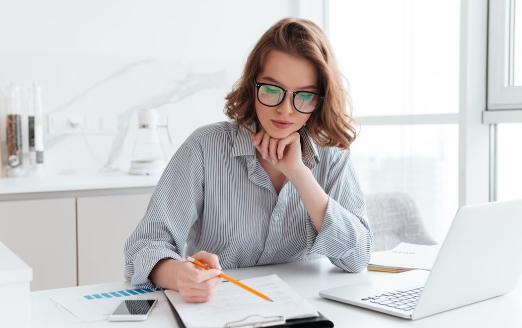 concentrated woman in glasses and striped shirt working with papers and laptop