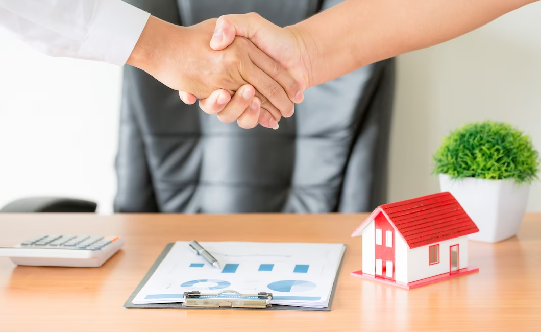 Two hands shaking above a table with a miniature mock building and documents