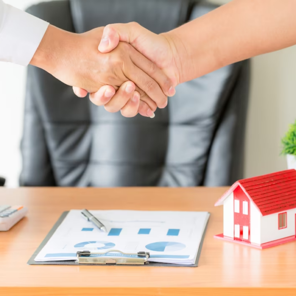 Two hands shaking above a table with a miniature mock building and documents