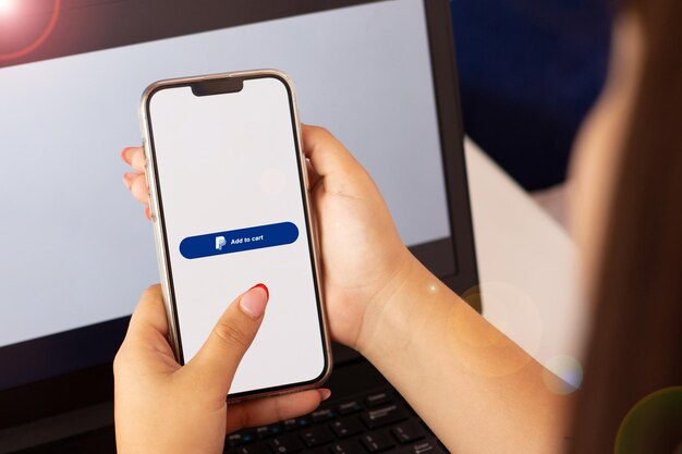 Hand holding a phone in front of a laptop displaying 'Add to Cart' option and a PayPal logo